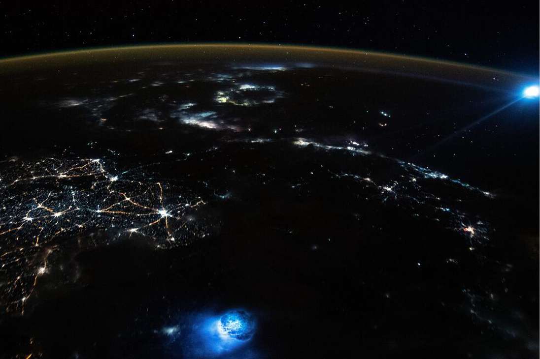 Un astronauta a bordo de la ISS tomó una imagen de la Tierra que contiene dos extrañas manchas azules de luz que brillan en la atmósfera de nuestro planeta. Curiosamente, las dos manchas no tienen ninguna relación entre sí y se han producido al mismo tiempo.
La primera mancha de luz, visible en la parte inferior de la imagen, es un enorme rayo que cayó junto a un gran hueco circular en la parte superior de las nubes.
La segunda mancha azul, que puede verse en la parte superior derecha de la imagen, es el resultado de la luz deformada procedente de la Luna.