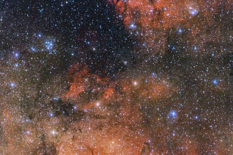 La imagen muestra un puñado de llamativas estrellas azules y nubes rojas de hidrógeno ionizado brillante y oscuros filamentos de polvo.  Los trazos oscuros que serpentean a través de la imagen son los turbios hilos de polvo cósmico que bloquean la luz de las estrellas distantes. / EFE 