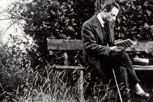 Rainer María Rilke, de impecable vestido, acompañado por tres objetos: a un lado, su sombrero, al otro, su bastón recostado en la pierna derecha, que está cruzada sobre su pierna izquierda, y en las manos; un libro, algo que nunca le podía faltar.  / Cortesía