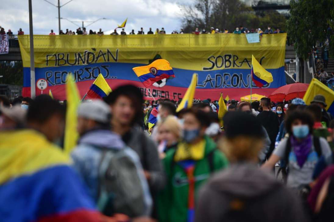 Varias personas caminando frente a una gran bandera de Colombia colgada en un puente.