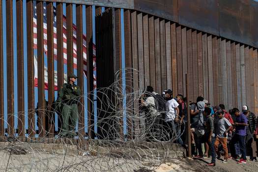 Migrantes centroamericanos miran a través de la frontera en el Chaparral (Baja California, México) hacia Estados Unidos, mientras un miembro de la patrulla fronteriza los vigila.  / AFP