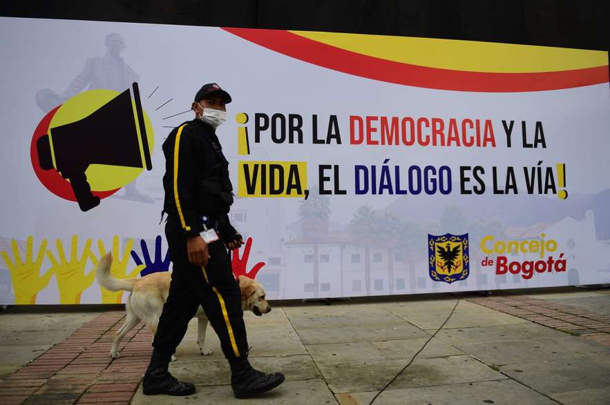Un celador con un perro pasando por el frente de un letrero que dice: Por la democracia y la vida, el diálogo es la vía
