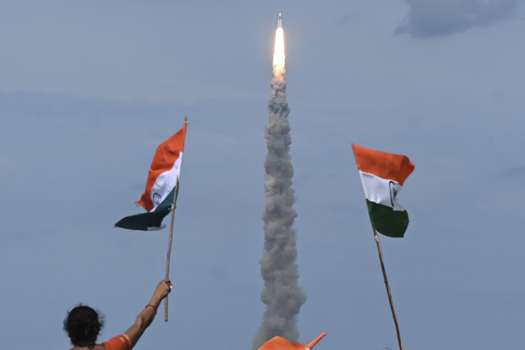 Varias personas ondean banderas indias mientras un cohete de la Organización India de Investigación Espacial (ISRO) que transporta la nave espacial Chandrayaan-3 despega del Centro Espacial Satish Dhawan de Sriharikota, una isla situada frente a la costa del estado meridional de Andhra Pradesh. (Foto por R.Satish BABU / AFP)