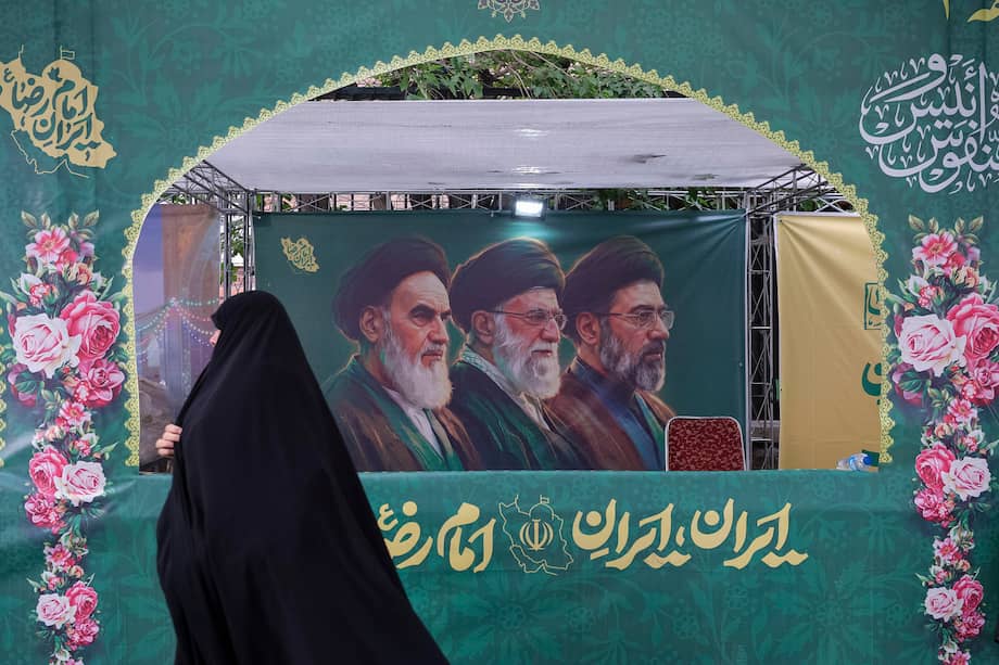 Una mujer vestida con el tradicional chador pasa delante de un cartel en Teherán que muestra a los tres líderes supremos de la República Islámica, incluido el actual Mojtaba Jameneí.