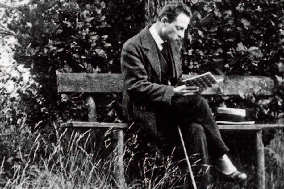 Rainer María Rilke, de impecable vestido, acompañado por tres objetos: a un lado, su sombrero, al otro, su bastón recostado en la pierna derecha, que está cruzada sobre su pierna izquierda, y en las manos; un libro, algo que nunca le podía faltar. / Cortesía