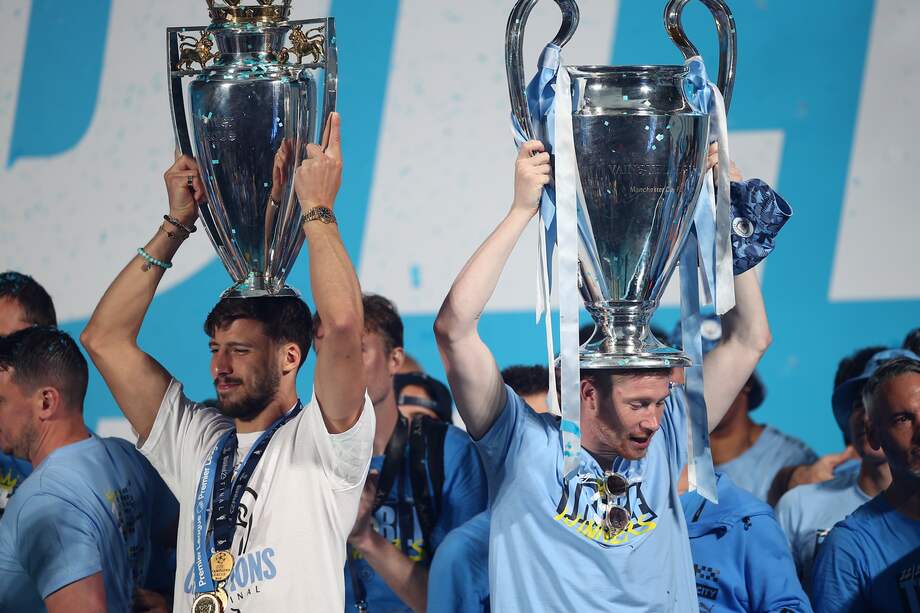 Ruben Dias (izquierda) y Kevin De Bruyne levantan los trofeos de la Premier League y de la Champions League en la celebración con sus hinchas en Manchester.