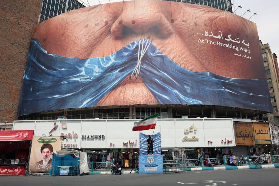 Un hombre sostiene la bandera nacional de Irán frente a una gran valla publicitaria antiestadounidense con la imagen del presidente Donald Trump y el estrecho de Ormuz.