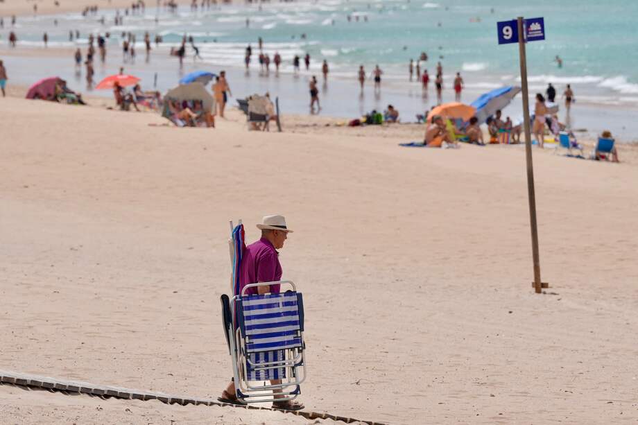 Un hombre cargado con sillas y sombrillas camina por la playa de Chiclana de la Frontera (Cádiz) en la que se ve al fondo numerosas personas, este viernes con temperaturas extremas de hasta 40 grados en Andalucía.