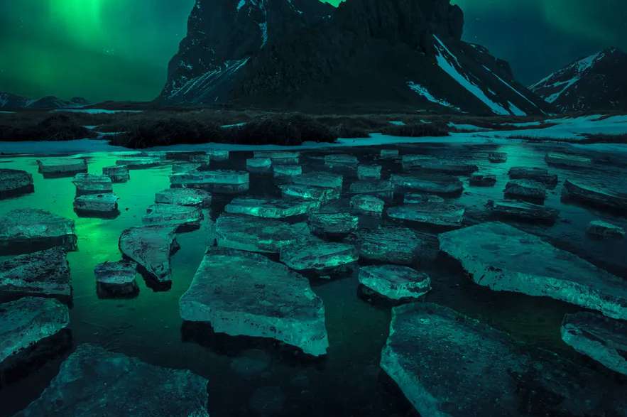 La imagen muestra la aurora boreal bailando, reflejada en un pequeño lago helado sobre la montaña Eystrahorn.