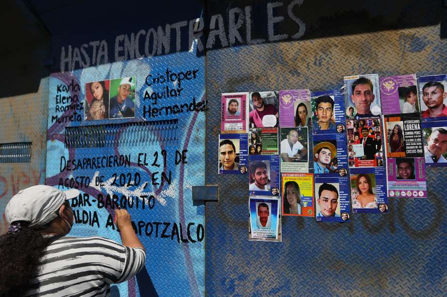 Una mujer escribe un mensaje en una valla junto a imágenes de personas desaparecidas.
