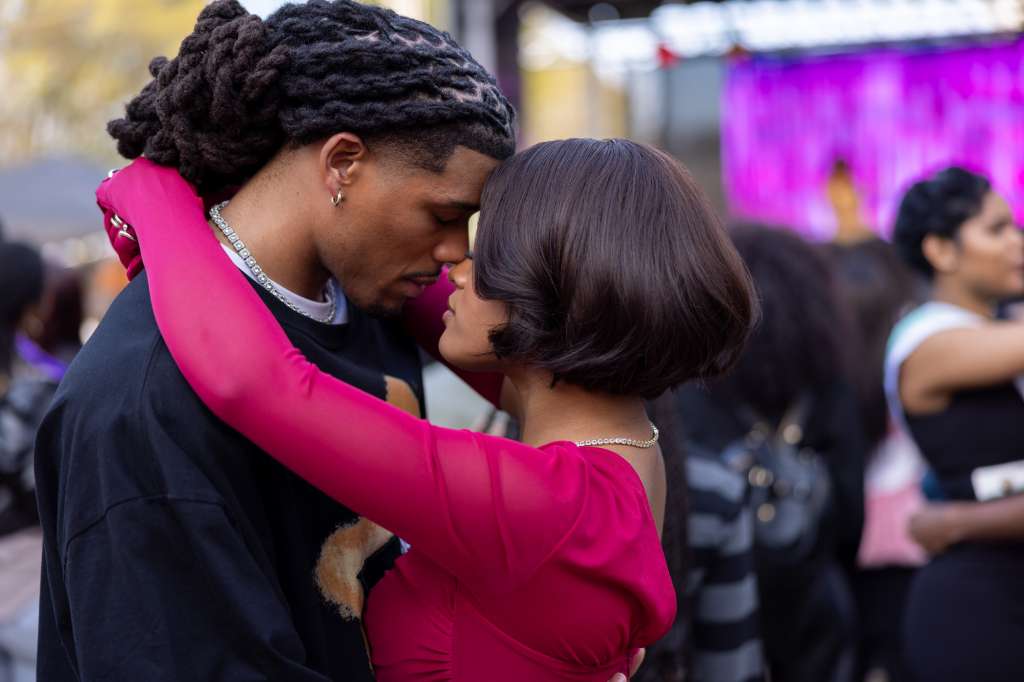 "A couple embraces closely in an outdoor public setting, their foreheads touching affectionately. Other people and a colorful stage are visible blurred in the background, suggesting a lively social event or festival."