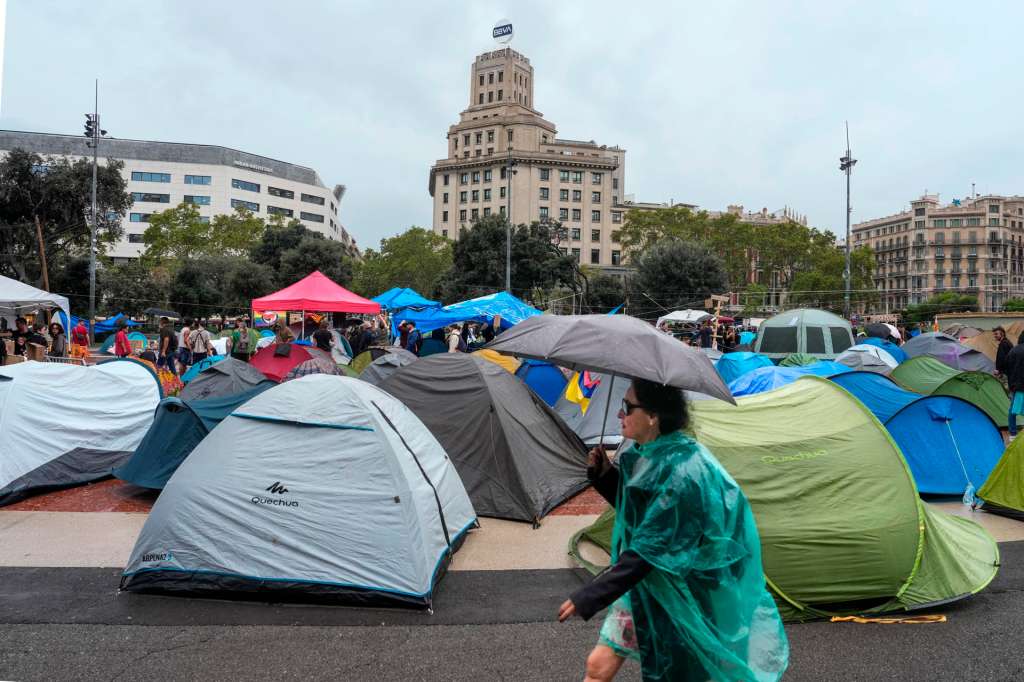 Una persona pasa con una sombrilla frente a la acampada instalada en la plaza de Catalunya de Barcelona.