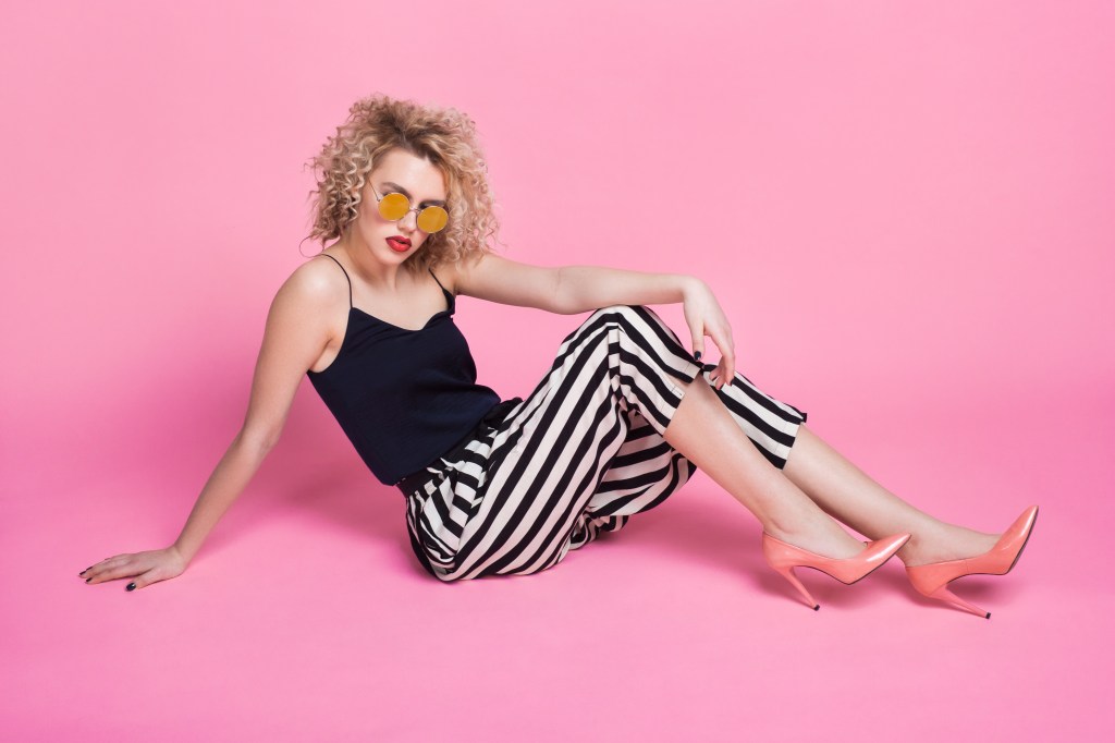 Young model in trendy outfit and sunglasses sitting and posing on pink.