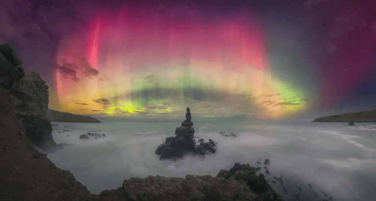 "Esta impresionante imagen panorámica, tomada durante una intensa tormenta G5, captura la vibrante aurora austral atravesando las nubes sobre una escarpada costa. Su fuerza reside en el equilibrio entre la precisión técnica y la resonancia emocional. La costa rocosa ancla la escena, contrastando con el brillo etéreo de la aurora. La maestría del fotógrafo queda patente en la perfecta unión de la panorámica y el refinado procesamiento del cielo. Tomada tras horas de esfuerzo, la imagen transmite tanto la belleza natural de la aurora como la dedicación del fotógrafo. Felicidades al fotógrafo por este notable logro".
