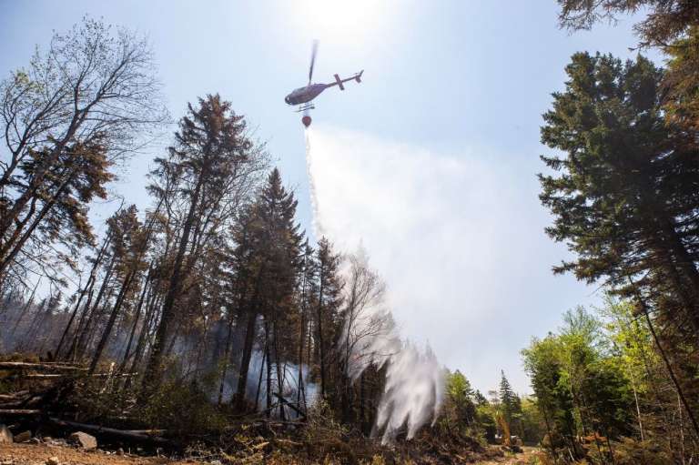 Esta imagen del 1 de junio de 2023, cortesía del Gobierno de Nueva Escocia en Canadá, muestra un helicóptero lanzando agua sobre un punto caliente detrás de Yankeetown Road mientras una excavadora hace un cortafuegos en el incendio forestal de Tantallon, Canadá.