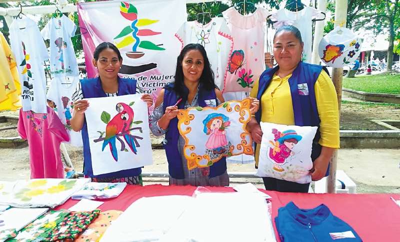 Tres mujeres de la Asociación de Mujeres Rurales y Víctimas de Tayarú muestran, sonrientes telares y cojines pintados a mano. Detrás de ellas hay camisetas y vestidos también estampados.