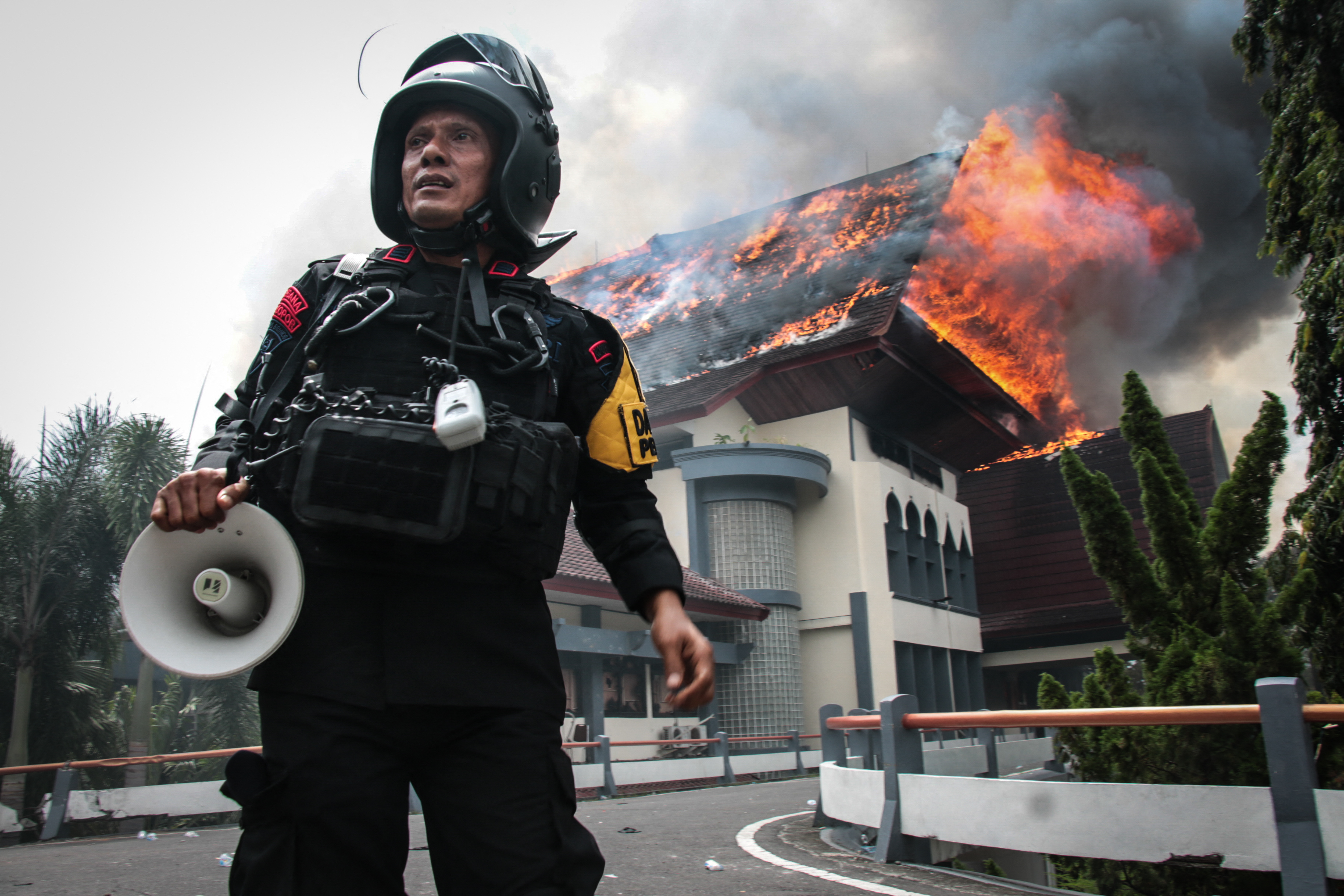 A police officer stands with a megaphone as the building of the West Nusa Tenggara Provincial Council office burns after being set on fire by protesters in Mataram on Lombok Island, West Nusa Tenggara, on August 30, 2025. Indonesia was rocked by protests in major cities including the capital Jakarta on August 29 after footage spread of a motorcycle taxi driver being run over by a police tactical vehicle in earlier rallies against low wages and financial perks for lawmakers. (Photo by Sadim / AFP)