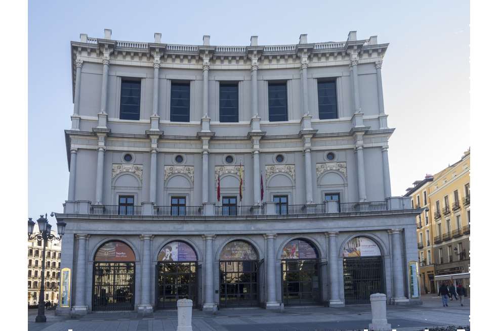 El Teatro Real de Madrid tiene una fachada de color blanco. Colgando de una de las ventanas del segundo piso, se ve la bandera de España.