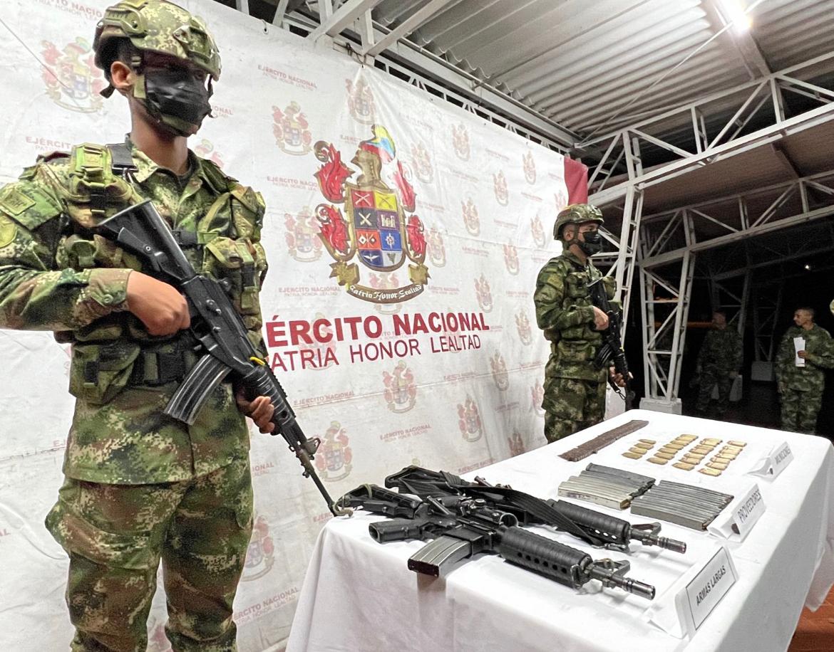 dos soldados con fusil, frente a una mesa con material de guerra incautado.