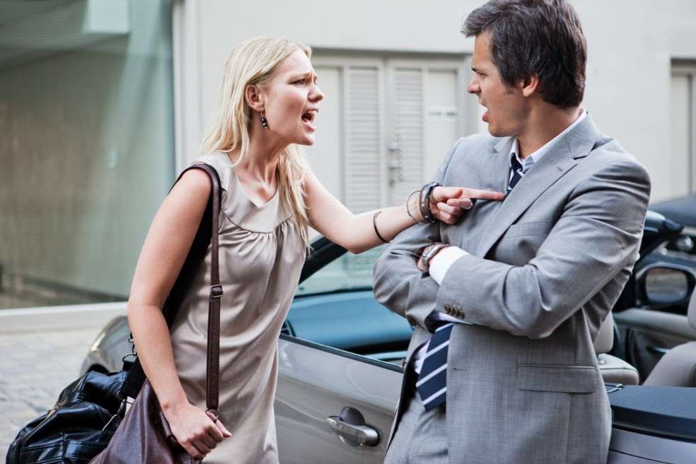 Una mujer rubia de vestido beige enterizo discute con un hombre de traje gris, camisa gris y corbata azul con líneas blancas diagonales, y que está recostado sobre un carro deportivo.