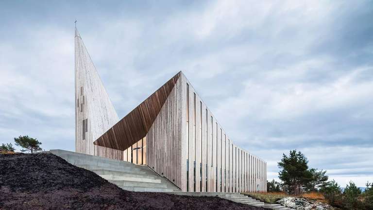 La Knarvik Church, diseñada por Reiulf Ramstad, es una interpretación contemporánea de las tradicionales iglesias de madera noruegas. Se encuentra en el municipio de Alver en el condado de Vestland, Noruega. Su estructura de líneas geométricas y una imponente aguja triangular revestida en pino envejecido se integra con el paisaje rocoso, mientras que en su interior los arcos y la luz natural crean un ambiente cálido y continuo. Inaugurada en 2014, no solo funciona como espacio religioso, sino también como centro cultural, conectando la herencia arquitectónica de Noruega con un diseño moderno y abierto a la comunidad.