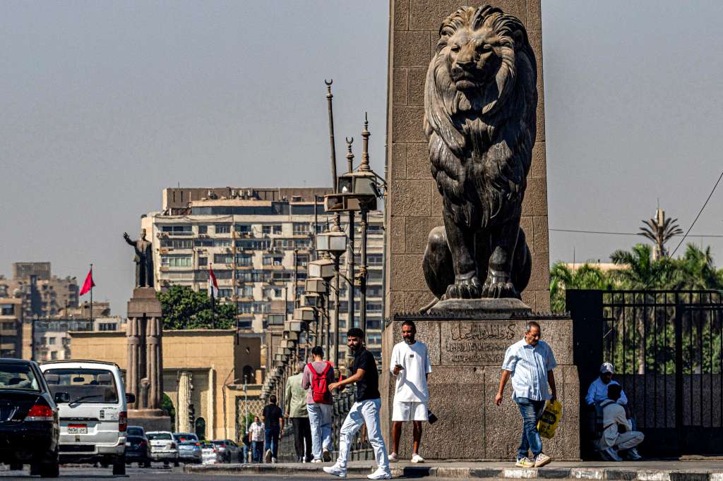 People walk past one of the four bronze lion statues at the end of the landmark Qasr el-Nile bridge connecting Cairo's central Tahrir Square to the island of Zamalek (background) on October 6, 2025.