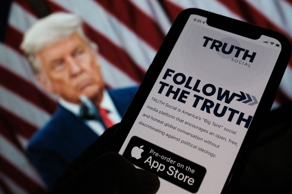 This illustration photo shows a person checking the app store on a smartphone for "Truth Social", with a photo of former US president Donald Trump on a computer screen in the background, in Los Angeles, October 20, 2021. - Former US president Donald Trump announced plans on October 20 to launch his own social networking platform called "TRUTH Social," which is expected to begin its beta launch for "invited guests" next month. The long-awaited platform will be owned by Trump Media & Technology Group (TMTG), which also intends to launch a subscription video on-demand service that will feature "non-woke" entertainment programming, the group said in a statement. (Photo by Chris DELMAS / AFP)
