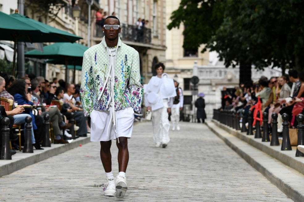 Un modelo luce una americana gris y camisa blanca que se combina con un foulard de seda rosa con motivos florales. Otro lleva un total lookcompuesto de camisa con hoodie y pantalón, recubierto con flores silvestres.