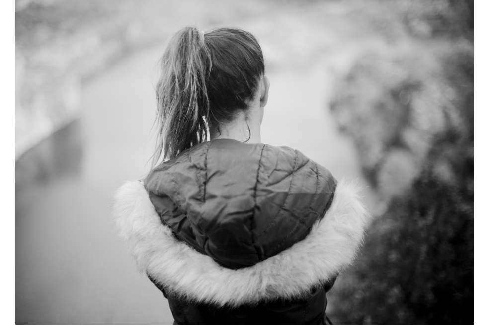 Fotografía a blanco y negro que muestra a una mujer de espaldas. Ella tiene puesta una chaqueta gruesa y el pelo cogido en una cola de caballo.