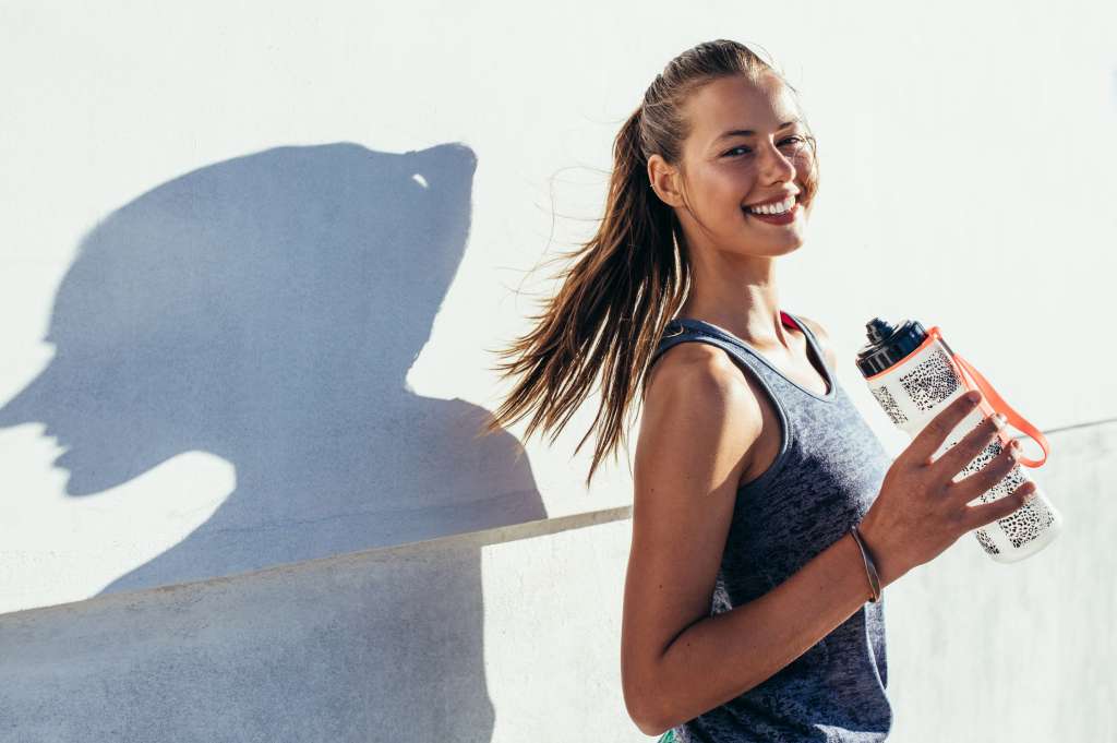 Shot of beautiful female runner standing outdoors holding water bottle. Fitness woman taking a break after running workout.