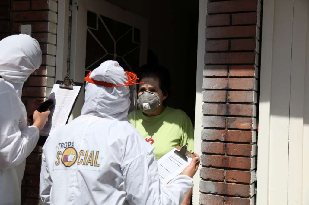 Dos personas con trajes blancos hablan con una mujer adulta, con tapabocas, en la entrada de una casa.