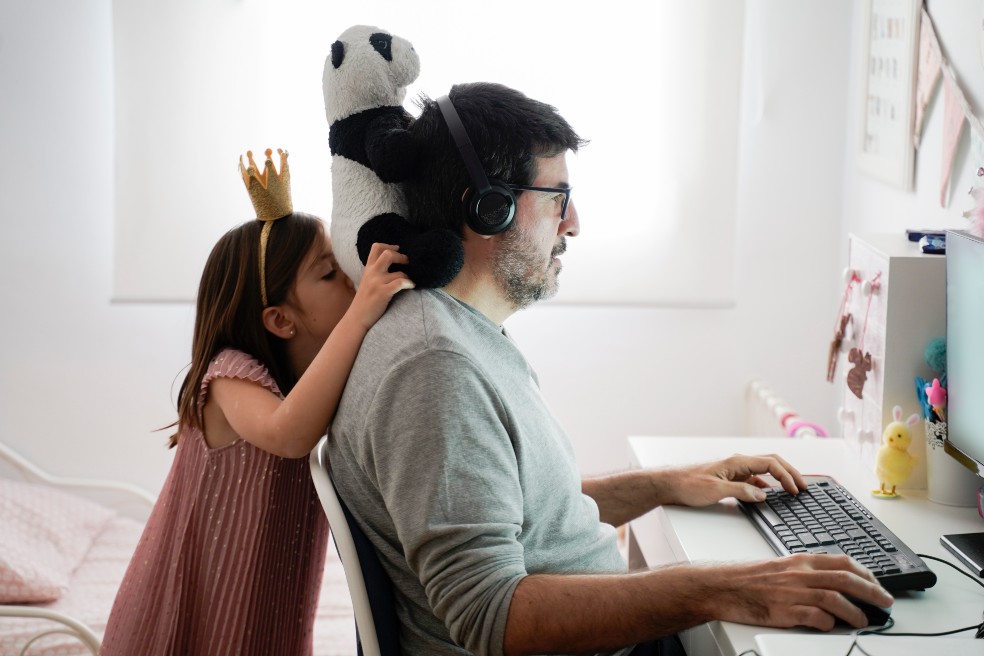 Un señor frente a un computador , mientras a sus espaldas su pequeña hija le pone un muñeco de trapo en la cabeza.