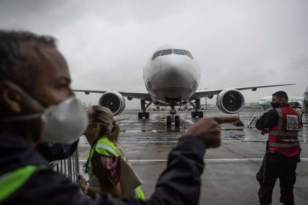 Un avión se ve en el foco de la imagen, pero en el fondo. En el frente hay personal de tierra del aeropuerto de Sao Paulo, Brasil, en sus labores, utilizando tapabocas.