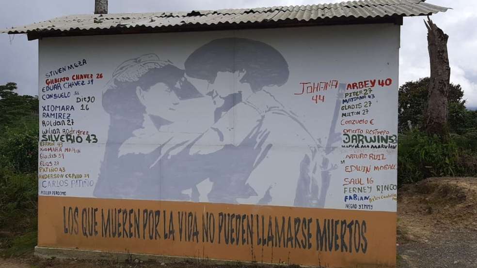 Una pared con con fondo blanco, tiene a sus laterales los nombres de personas excombatientes asesinadas.