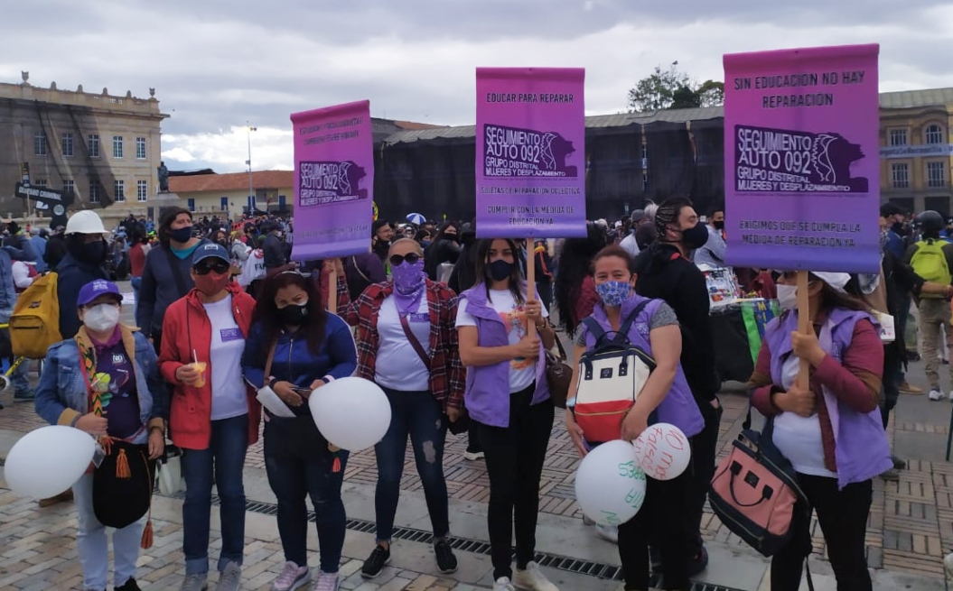 Un grupo de siete mujeres se manifiestan en la plaza de Bolívar, detrás de ellas se ven muchos otros ciudadanos. Tienen tapabocas y portan carteles que dicen "Sin educación no hay reparación".