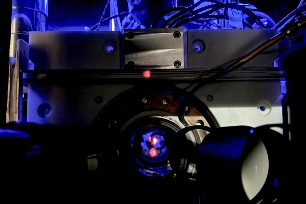 Un gas extremadamente frío de átomos de estroncio está atrapado en una red de luz conocida como red óptica. Los átomos se mantienen en un entorno de vacío ultraalto, lo que significa que casi no hay aire ni otros gases presentes. Este vacío ayuda a preservar los delicados estados cuánticos de los átomos, que son frágiles. El punto rojo que se ve en la imagen es un reflejo de la luz láser utilizada para crear la trampa de átomos.