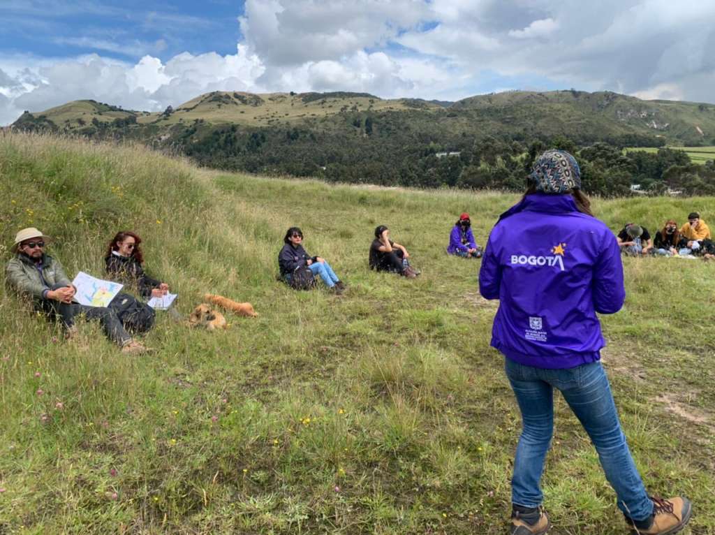 Una mujer de espaldas, frente a otras siete que están sentadas en una zona verde.