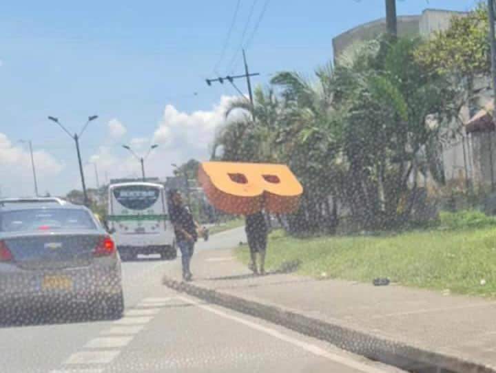 Hombre, quien se esta llevando la letra B hombro arriba, camina tranquilamente por una vía transitada.