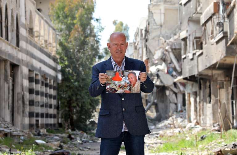 Mohammed al-Rakouia, un pintor refugiado palestino de 70 años del devastado campo de Yarmouk para refugiados palestinos al sur de la capital de Siria, Damasco, se encuentra junto a un callejón junto a los edificios dañados en el campo el 7 de marzo de 2021, mientras sostiene una foto de sí mismo. trabajando en su antiguo estudio que data de diez años antes. Al-Rakouia lamenta sus pérdidas diciendo que "nada puede compensarlas". "Mi estudio ha sido destruido, mis pinturas han sido robadas y mis colores han sido esparcidos por todo el lugar".