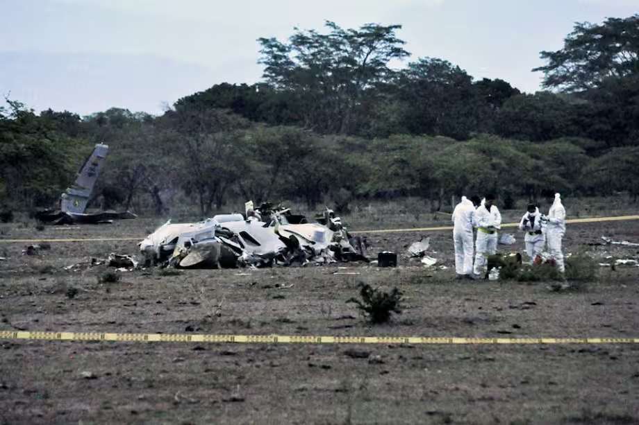 Restos de una aeronave militar en un campo abierto, con fuselaje destruido y la cola parcialmente en pie. Personas con trajes blancos de protección inspeccionan el lugar, rodeado por cinta amarilla de seguridad.
