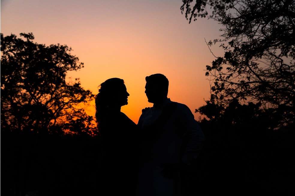 Pareja en el atardecer. La luz naranja del sol sale en el centro de la foto, mientras las sombras de la pareja y de los árboles se ven a su alrededor.