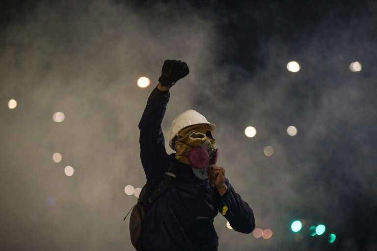 Un hombre con casco y mascara antigas levantando el brazo derecho con el puño cerrado.