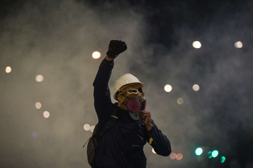 Un hombre con casco y mascara antigas levantando el brazo derecho con el puño cerrado.
