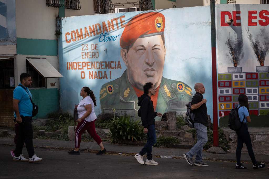 AME9700. CARACAS (VENEZUELA), 18/03/2024.- Varias personas caminan frente a un mural del fallecido presidente venezolano Hugo Chávez este lunes, en Caracas (Venezuela). A un mes para que venza el plazo de seis meses establecido por Estados Unidos para la suspensión de algunas sanciones a Venezuela, entre ellas al petróleo y al gas, se eleva el suspenso respecto a la decisión que, a la postre, tome el Gobierno estadounidense, que supedita la extensión del alivio a los pasos que dé Caracas para garantizar en julio unas presidenciales "competitivas". EFE/ Rayner Peña R.