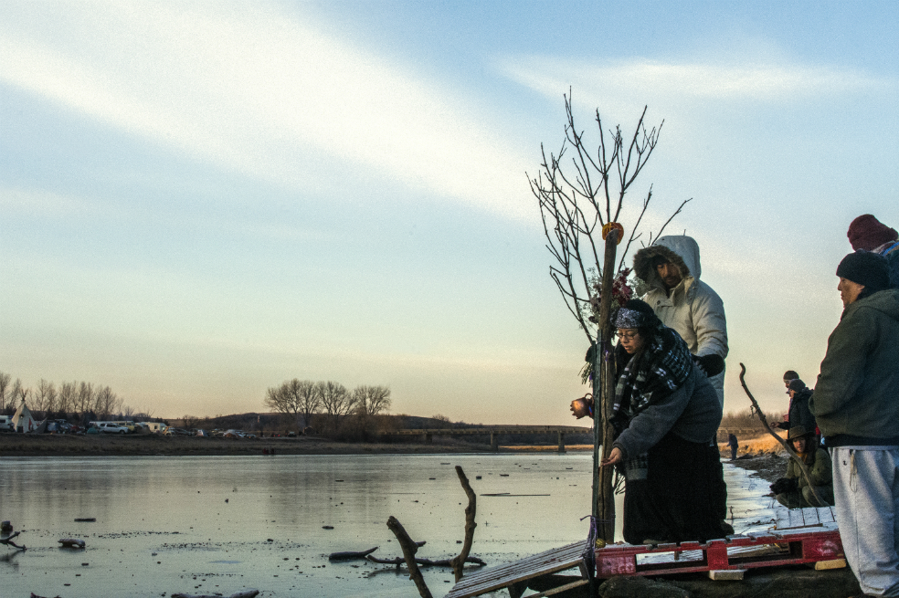 Protectores del Agua en ceremonía sagrada por el río Missouri en el duro invierno de Dakota del Norte. La mujer echa tabaco al río y lo bendice con agua que riega sobre él. Campamento Oceti Sakowin, Dakota del Norte. / Alexandra McNichols-Torroledo