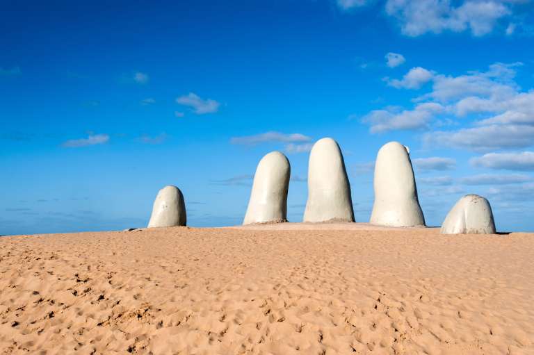 Escultura de los dedos en Uruguay
Cuenta la leyenda que los deseos de aquellos enamorados que se besen sobre la mano de Playa Mansa se cumplirán si son sinceros. Ubicada en la costa de Río de la Plata, Maldonado, se ha convertido en unos de los principales puntos de interés para aquellos que buscan una escapada tranquila y romántica entre arena fina y olas de pequeño tamaño.
De camino a la mano bajo la playa, es parada obligatoria el Faro, la construcción más antigua de la península , los enormes nidos de amor de San Rafael y los característicos puentes ondulantes.