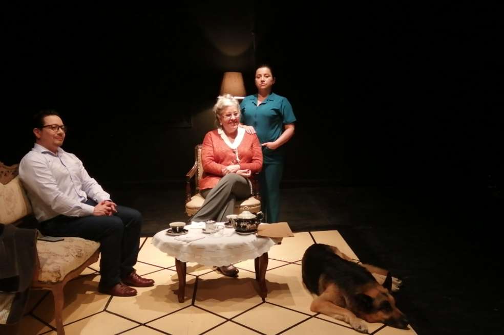 En escena hay cuatro personajes de la obra "Si me muero es suya". Uno de ellos, vestido con camisa clara, está sentado en un sofá. La protagonista está vestida con un saco rosado y sentada en una silla. A su lado hay una mujer vestida con uniforme verde. En el piso, acostado, hay un perro.