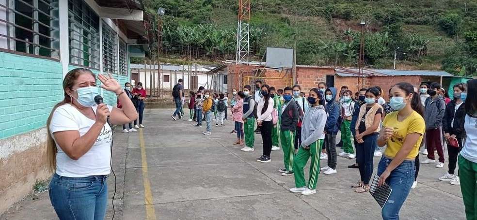 Una profesora, de camiseta blanca u blue jean, les habla a varios alumnos adolescentes en el patio de un colegio, en el Catatumbo, subregión de Norte de Santander.