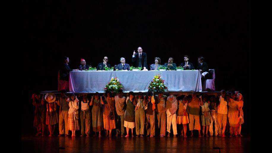 Escena "El banquete" en el que se representa a sectores políticos y un sector del país que justifican la guerra desde sus escritorios, en palabras de Andrés Parra (en el centro de la imagen).