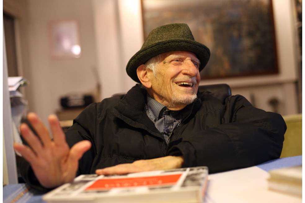 Nereo López está sentado, vestido con ropa oscura y un sombrero. Está sonriendo.